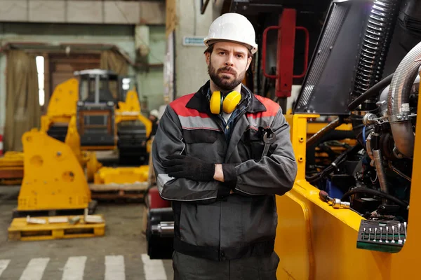 Young serious worker of plant in hardhat and uniform standing by huge industrial machine and keeping his arms crossed on chest