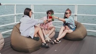 Tilting down of two girls and guy sitting on pier on lake on hot summer day, drinking beer and cocktails, talking and smiling