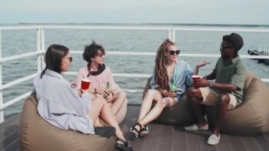 Slow motion of African American guy greeting friends of his Caucasian girlfriend, sitting on pier on lake on hot summer day, drinking beer and cocktails, shaking hands talking and smiling