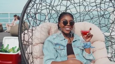 Medium portrait of delighted African American girl wearing sunglasses, sitting in hanging chair, holding glass with bright cocktail, smiling on camera