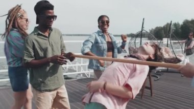 Medium long of young friends having party on lake pier, playing limbo, laughing and cheering on hot summer day