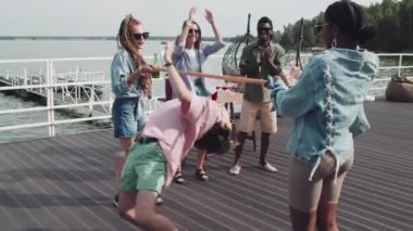 Young friends having party on lake pier, playing limbo, drinking beer, laughing and clapping hands on hot summer day