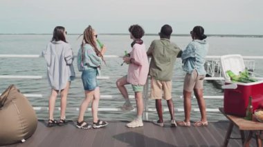 Rear of five multiethnic girls and guys standing on edge of pier on lake, drinking beer, chatting and looking at water on sunny summer day