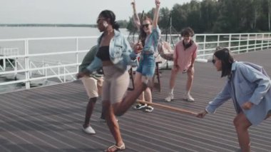 Medium long of young friends having party on lake pier, playing with vertical bar, jumping, laughing and high-fiving on hot summer day