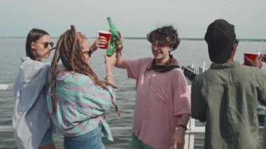Medium long of multiethnic girls and guys standing on edge of pier on lake, clinking bottles and plastic cups, drinking beer and beverages on sunny summer day