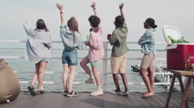 Rear of five multiethnic girls and guys standing on edge of pier on lake, drinking beer, waving hands and looking at water on sunny summer day