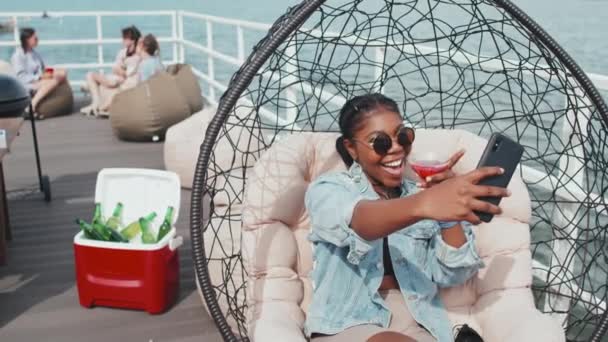 喜んでいるアフリカ系アメリカ人の女の子の中には サングラスをかけ 椅子に座り 明るいカクテルを片手にガラスを持ち スマートフォンで自撮り — ストック動画