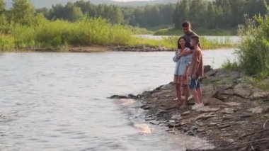 Wide shot of Caucasian boy with young mother and father standing on rocky coast of lake on sunny summer day, talking
