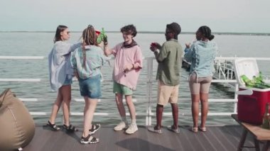 Rear of five multiethnic girls and guys standing on edge of pier on lake, looking at water, then clinking bottles and plastic cups, drinking beer and beverages on sunny summer day