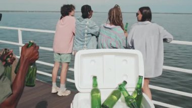 Slow motion of young multiethnic female and male friends in sunglasses and summer clothes having fun and drinking on lake pier on hot day