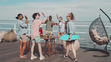 Tracking of Caucasian girl with dreadlocks carrying skateboard, walking on pier on lake, greeting young multiethnic friends, celebrating holiday in summer heat