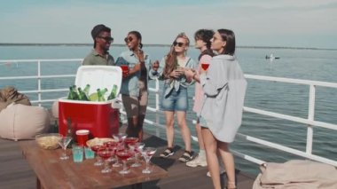 Slow motion of young multiethnic female and male friends in sunglasses and summer clothes dancing, having fun and drinking on lake pier on hot day