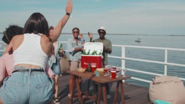 Biracial guy carrying dark-haired girl on his back, couple having fun on hot summer weekend, diverse young friends watching them, standing on pier on lake