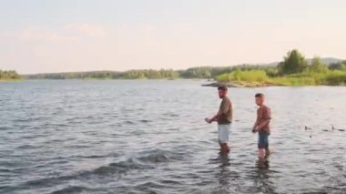 Side view of young Biracial man and teenage boy wearing shorts and tees, standing in lake, throwing stones in water on warm summer day
