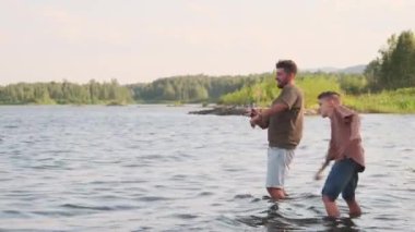 Side view of young Biracial man and teenage boy wearing shorts and tees, standing in lake, throwing rocks on warm summer day