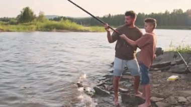 Full long side view of young father teaching teenage son who wearing shorts and tees, fishing, standing on rocky on lake shore, talking
