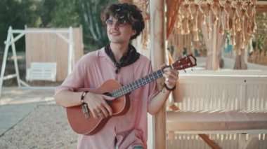 Medium of dark-haired Caucasian guy in sunglasses playing ukulele, standing by gazebo on beach, smiling on hot summer day
