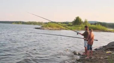 Long side view of young Biracial man and teenage boy wearing shorts and tees, standing on rocky on lake shore, chatting and fishing