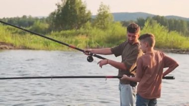 Medium long side view of young father and teenage boy wearing shorts and tees, standing on rocky on lake shore, talking and fishing