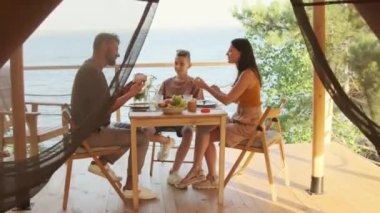 Full shot of young happy woman and man and teenage boy sitting at table on terrace by lake on sunny summer day, having breakfast