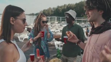 Medium of multiethnic girls and guys having snacks and drinks, chatting, having fun on beach on summer holiday