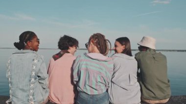 Rear medium of five multiethnic girls and guys wearing summer clothes, talking, sitting on lakeshore on hot and sunny day