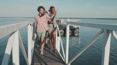 Group of diverse young friends walking on footbridge on pier on lake in summer, talking and laughing