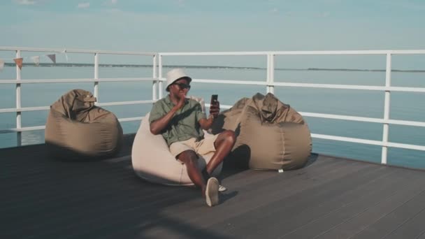 Wide Shot Young Black Man Sitting Bean Chair Pier Lake — Vídeos de Stock