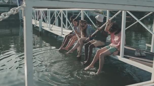 夏の服を着た5人の多民族の女の子と男の広いショット 湖の桟橋の歩道橋の上に座って 友人は暑い晴れた日の足を濡らす — ストック動画