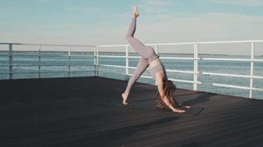 Wide of young Caucasian woman with dreadlocks wearing matching leggings and sports top, doing yoga poses on pier by lake in summer morning, stretching