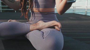 Low angle of cropped Caucasian woman with dreadlocks wearing matching leggings and sports top, sitting in lotus pose on yoga mat on pier by lake in summer morning, meditating