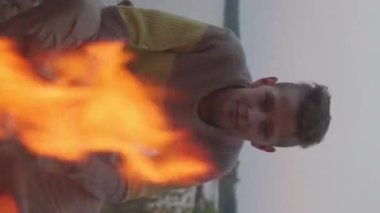 Vertical of teenage Caucasian boy sitting on lakeshore, watching campfire in summer evening