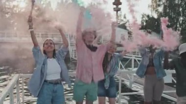 Medium long of diverse young people standing with colorful smoke bombs on pier by lake in summer evening