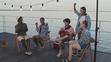 Wide shot of company of young diverse people sitting on pier by lake in summer evening, Caucasian guy playing ukulele and friends singing along