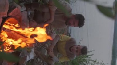 Vertical of teenage Caucasian boy sitting on lakeshore, watching campfire in summer evening, talking with young father