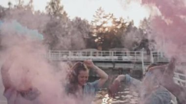 Medium of diverse young people standing with colorful smoke bombs on pier by lake in summer evening