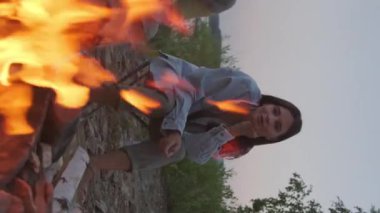 Vertical of young dark-haired Biracial woman sitting on lakeshore, watching campfire in summer evening, talking