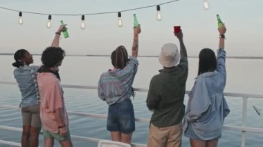 Rear medium long of five diverse young people holding drinks up, standing by fencing, looking at lake in summer evening, celebrating holidays