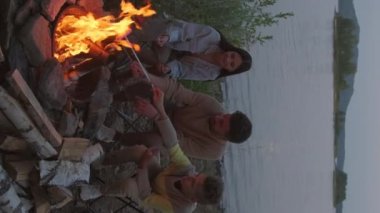 Vertical long of happy family sitting on lakeshore by campfire on summer evening, roasting sausages on sticks, talking and enjoying nature