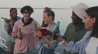 Medium of five diverse authentic young friends clinking beer bottles, laughing, standing on pier on lake in summer evening, drinking and partying
