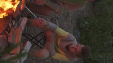 Vertical of teenage Caucasian boy and cropped man sitting on lakeshore by campfire on summer evening, roasting sausages on sticks, talking and enjoying nature