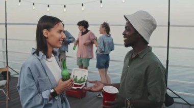 Waist-up of Biracial girl and Black guy having alcoholic drinks, talking, standing on pier by lake, celebrating summer vacation party in evening