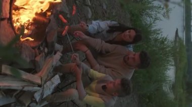 Vertical of young bearded Biracial man sitting with young wife and teenage son on lakeshore by campfire on summer evening, roasting sausages on skewers, talking and enjoying nature