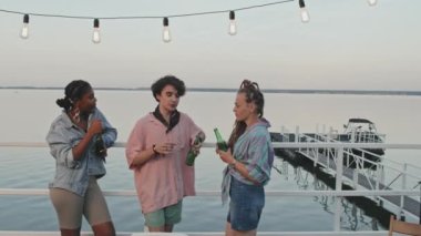 Medium long of diverse guy and two girls having beer, talking, standing on pier by lake, celebrating summer vacation party in evening