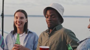 Chest-up of young Black man talking to diverse friends, standing outdoors, drinking, laughing, having lake party in summer evening