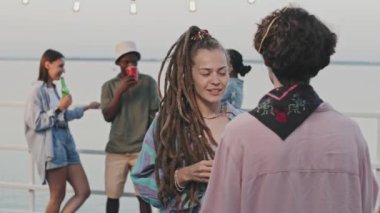 Waist-up slow motion of authentic girl with dreadlocks drinking beer from bottle, talking to Caucasian guy, standing on pier in warm summer evening