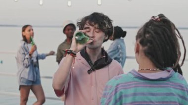 Waist-up of Caucasian guy drinking beer from bottle, talking to authentic girl with dreadlocks, standing on pier in warm summer evening