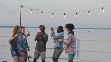 Medium long of energetic diverse young friends dancing and drinking on lake party in summer evening