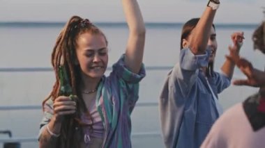Medium close-up of authentic multiethnic young friends raising hands with drinks up in air, dancing and having fun on party in nature in summer evening