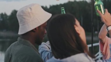 Medium close-up of cheerful multiethnic young friends raising hands with drinks up in air, dancing and smiling on lake party in summer evening
